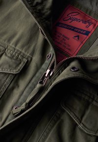 Khaki zielona kurtka robocza o strukturalnym kształcie, metalowy zamek błyskawiczny oraz czerwona metka z informacjami o marce i instrukcjami pielęgnacyjnymi.