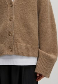 Bruine gebreide cardigan met een losse pasvorm, voorzien van grote knopen en geribbelde manchetten. Gemaakt over een wit shirt en een zwarte rok. Zachte textuur.