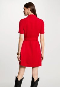Vestido rojo de manga corta con cintura ajustada y cinturón, que presenta cuello de solapa y textura suave. Botas de cuero marrón visibles en la parte inferior.