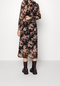 Kvinna som står med ryggen vänd mot betraktaren, iförd en svart blommig midiklänning med långa ärmar och mörkbruna ankla stövlar.