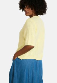 Īsām piedurknēm dzeltena adīta džempera ar teksturētu rakstu un nedaudz saīsinātu piegriezumu, apvienots ar zilu džinsu svārku.