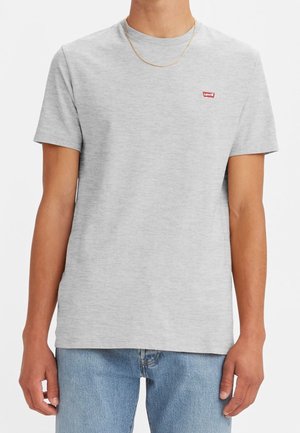 Osoba nosząca jednolity jasnoszary t-shirt z krótkim rękawem, z małym czerwonym logo Levi's na klatce piersiowej i jasnoniebieskie dżinsy.