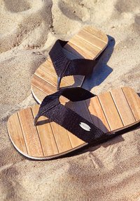 Wooden flip-flops with black woven straps and silver accents, laid on sand, showcasing a slatted wooden footbed.
