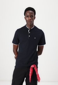 Dunkelblauses Polohemd mit Knopfverschluss und weißen Kragenakzenten, kombiniert mit schwarzen Cargo-Shorts und einem roten Halstuch, das um die Taille gebunden ist.