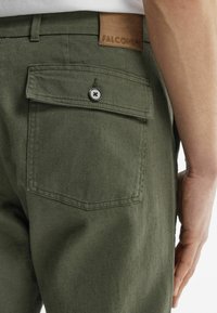 Olivgröna byxor i en texturerad tygkvalitet, med en bakficka med knappstängning och en kontrasterande brun läderlapp som är märkt "FALCONERI."