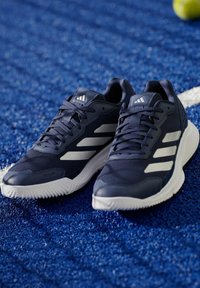 Двойка тъмносини спортни обувки Adidas с бели подметки и райета върху синя текстурирана повърхност с частично видима тенис топка.