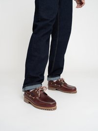 Chaussons de bateau en cuir marron avec des lacets beige, bout arrondi et détails de couture visibles. Portés avec un jean en denim foncé et des revers.