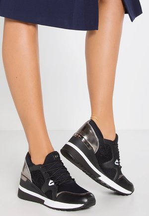 Jambes portant des baskets compensées noires et métalliques avec des semelles intermédiaires blanches, associées à une jupe bleu marine longueur genou, debout sur un sol blanc.