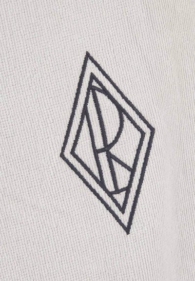 Μαύρο γεωμετρικό λογότυπο με επικαλυπτόμενα γράμματα «A» και «R» μέσα σε σχήμα ρόμβου, κεντημένο σε υφασμάτινο φόντο με υφή λευκού χρώματος.