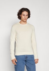Light beige knit sweater with long sleeves, ribbed cuffs, and neckline, paired with blue denim jeans. Smooth texture, fitted design.
