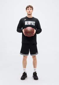 Μαύρο πουκάμισο με μακριά μανίκια και το κείμενο «MEMPHIS», μαύρο σορτς με λευκές ρίγες, κρατώντας μια κόκκινη μπάλα μπάσκετ Spalding, φορεμένο με μαύρα αθλητικά παπούτσια.