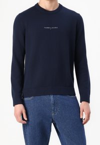 Granatowy sweter dzianinowy z prążkowanym okrągłym dekoltem, z białym logo na piersi oraz prążkowanymi mankietami i dołem. Połączony z niebieskimi dżinsami.