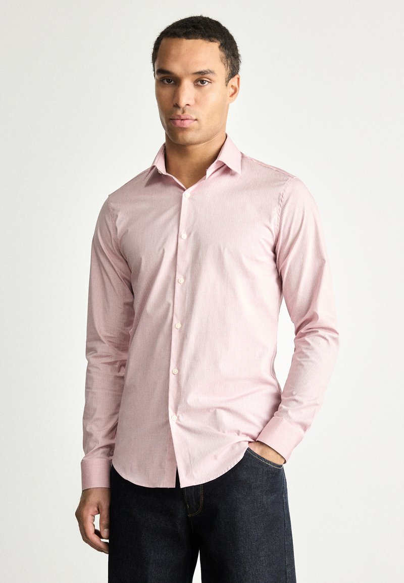 Chemise rose clair avec de fines rayures verticales, manches longues, un col classique et des boutons blancs, associée à un jean en denim bleu foncé.