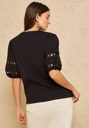 Nainen mustassa paidassa, jossa on silmäreikäkaitaleilla varustetut pussihihat, yhdistettynä korkeavyötäröisiin valkoisiin housuihin, seisomassa teksturoidun seinän edessä.