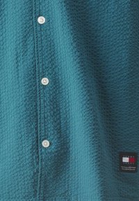 Camicia teal con bottoni, realizzata in tessuto texturizzato. Presenta bottoni bianchi e un'etichetta rettangolare con il logo del marchio in basso a destra.