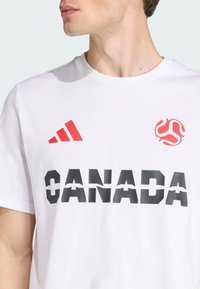 Camiseta de algodón blanca con un gráfico negro que dice "CANADÁ" y el logo y detalles en rojo de Adidas. Cuello redondo, mangas cortas y textura suave.