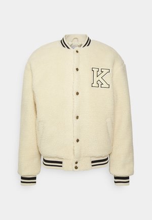 Chaqueta varsity de forro polar color crema con cierre de botones, puños y cuello a rayas en blanco y negro, y parche con la letra "K" en el pecho.