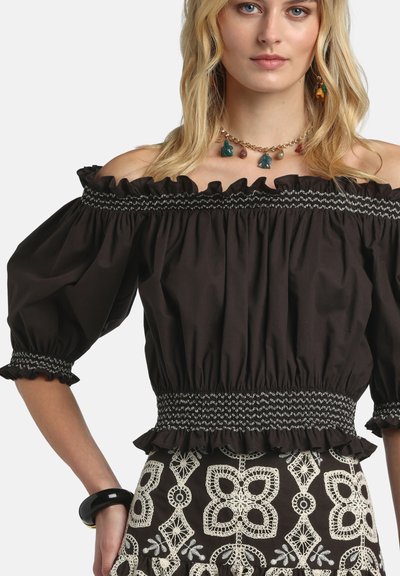 Femme portant un chemisier noir à épaules dénudées avec des bords à volants et une jupe à motifs, accessoirisée d'un collier porte-bonheur coloré et d'un bracelet noir.