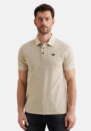 Hombre con camiseta tipo polo beige con patrón, cuello y logo de mariposa en el lado izquierdo del pecho, de pie contra un fondo blanco.