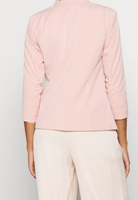 Blazer rose pâle ajusté avec un col structuré, des manches aux trois quarts, et une texture de tissu lisse, associé à un pantalon large de couleur claire.