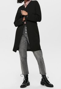 Czarny płaszcz z długimi rękawami, noszony na jasnoróżowej koszuli z kołnierzykiem, w zestawieniu z szarymi dżinsami i czarnymi sznurowanymi botkami. Gładka tekstura.