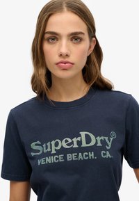 Marineblå bomulds t-shirt med korte ærmer, prydet med et stort, prikket "SuperDry" logo og teksten "Venice Beach, CA" i kontrasterende farver.