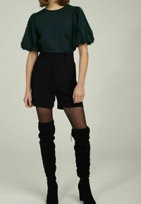 Haut vert foncé à manches courtes et manches bouffantes ; short noir taille haute ; collants noirs à pois ; bottes noires en daim montant jusqu'aux genoux.