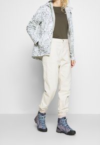 Vaellusasu, joka sisältää kevyen kukallisen takin, oliivinvihreän paidan, kermaiset cargo-housut ja siniset vaelluskenkä, joissa on harmaita ja mustia yksityiskohtia.