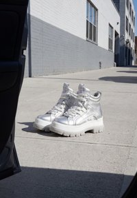 Bottes plateformes épaisses argentées avec semelles blanches épaisses placées sur un trottoir en ville près d’un bâtiment en briques gris et blanc.