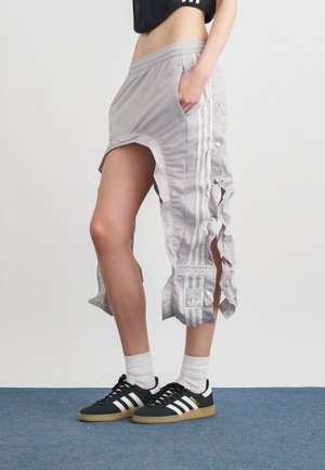 Kobieta w czarnym krótkim topie, jasnoszarych spodniach Adidas zapinanych z boków, białych skarpetkach oraz czarnych butach sportowych Adidas na niebieskiej podłodze.