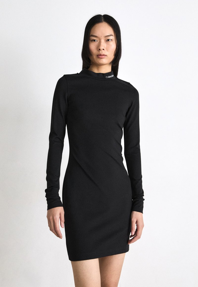 Long-sleeve black dress made of smooth fabric, featuring a high neck and a small logo detail at the collar. Fitted silhouette and above-knee length.