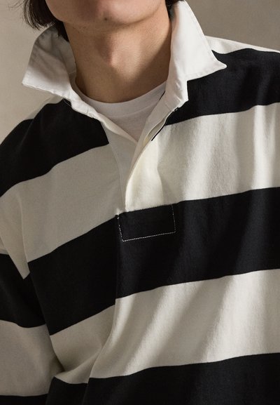 Persoon draagt een zwart-wit horizontaal gestreept rugbyshirt met een open witte kraag over een wit T-shirt met ronde hals.