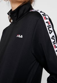 Veste noire zippée en tissu lisse. Caractérisée par un col montant et des bandes latérales blanches contrastantes avec du texte rouge et noir. Logo Fila sur la poitrine.