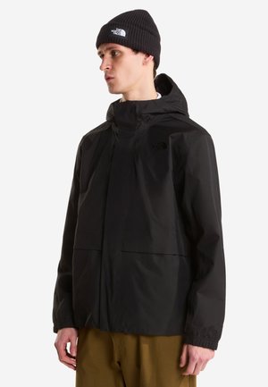 Jeune homme portant une veste imperméable noire avec capuche et un bonnet en laine noir affichant le logo The North Face.