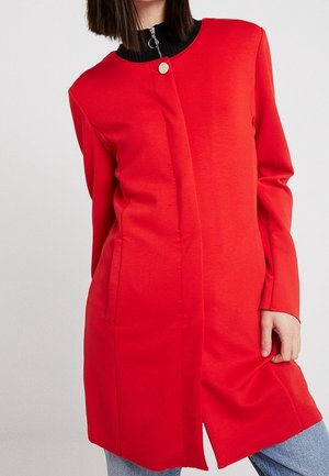 Femme portant un long manteau rouge sur un haut noir et un jean bleu, une main tenant le bord du manteau.