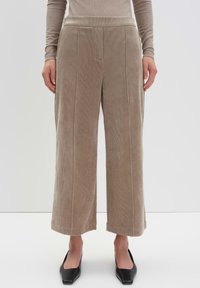 Pantalons en velours côtelé beige à jambes larges et raccourcies portés avec un haut à manches longues et des chaussures noires à bout pointu sur fond blanc.
