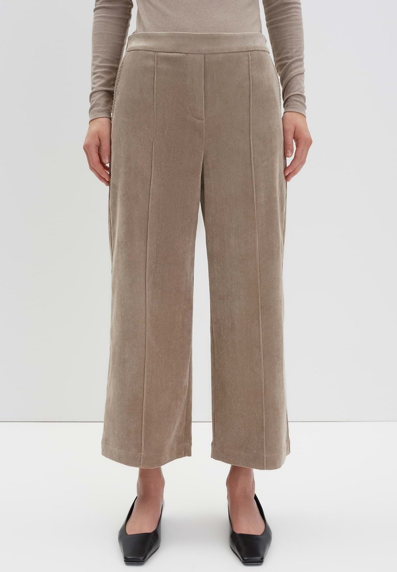 Pantalons en velours côtelé beige à jambes larges et raccourcies portés avec un haut à manches longues et des chaussures noires à bout pointu sur fond blanc.