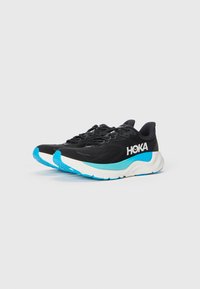 Schwarze Sportschuhe mit einem atmungsaktiven Obermaterial, ausgestattet mit einer weißen und türkisfarbenen Sohle sowie Branding. Strukturierte Materialien verbessern den Halt und den Komfort.