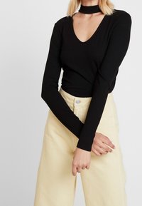 Musta ribattava pitkähihainen paita, jossa on syvä V-pääntie ja choker-yksityiskohta, yhdistettynä vaaleankeltaisiin korkeavyötäröisiin leveälahkeisiin housuihin.