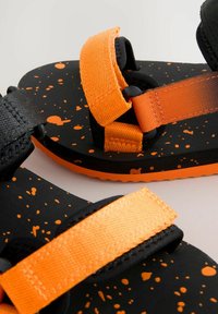 Mustangit musta sandaalit oranssilla roiskepinnoitteella, joissa on säädettävät oranssit hihnat ja teksturoitu musta materiaali, joka tarjoaa seuraavan tason pitoa ja mukavuutta.