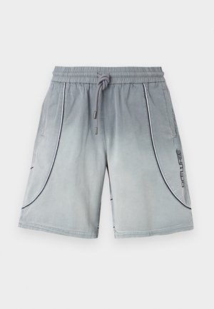 Mann trägt schwarze Sonnenbrille, dunkelblauen und hellblauen Pullover mit der Zahl "00" vorne, graue Shorts, schwarze Socken und schwarz-weiße Turnschuhe, steht vor einer weißen Wand.