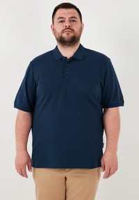 Tamsiai mėlynas polo marškinėliai, pagaminti iš medvilnės mišinio, su apykakle, trumpomis rankovėmis ir trimis mygtukais. Lygus tekstūra, be raštų ar akcentų.