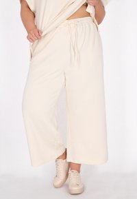 Cremefarbene, weit geschnittene Hose mit einem strukturierten diagonalen Muster und elastischem Bund, kombiniert mit hellbeigen Sneakers.