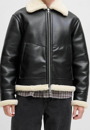 Veste en cuir noire avec col, poignets et bas en peau lainée blanche portée sur une chemise à carreaux et un jean foncé.
