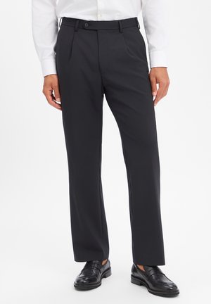 Mann trägt schwarze, maßgeschneiderte Hose mit Seitentaschen, weißes Hemd und schwarze Leder-Loafer vor weißem Hintergrund.