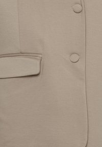 Beige blézer texturált anyagból, látható zsebbel, két kerek gombbal és sima felületű, testhezálló fazonnal.
