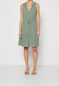 Abito verde corto senza maniche con scollatura a V, collo arricciato, vita regolabile con cordoncino e gonna a strati. Indossato con sandali beige a scivolo.