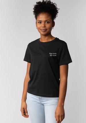 Schwarz Baumwoll-T-Shirt mit kurzen Ärmeln und rundem Ausschnitt. Mit weißem Text auf der linken Brust, der "Was muss, das muss." lautet.