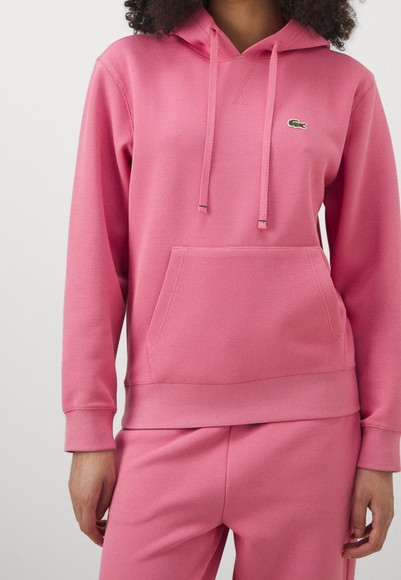 Sweat à capuche rose en tissu texturé avec une poche avant, des cordons de serrage et un petit logo brodé sur le côté gauche de la poitrine. Pantalon rose assorti.