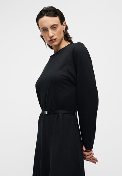 Μαύρο φόρεμα με μακριά μανίκια και εφαρμοστή μέση, τονισμένη από λεπτή μαύρη ζώνη με ασημένια αγκράφα, με ομαλή, ματ υφή.